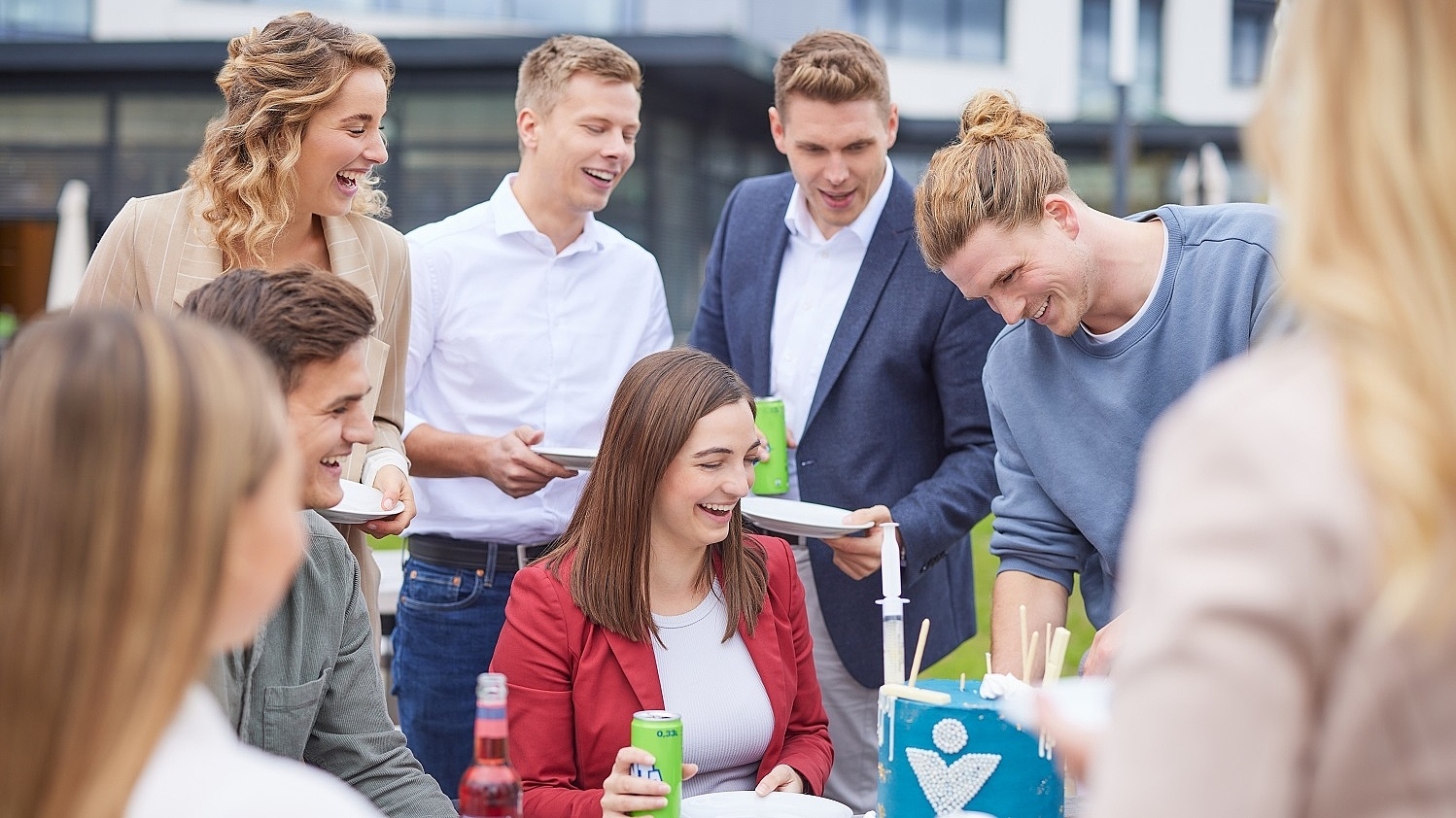 Lächelnde Mitarbeiter stehen und sitzen um einen Tisch und essen Kuchen 