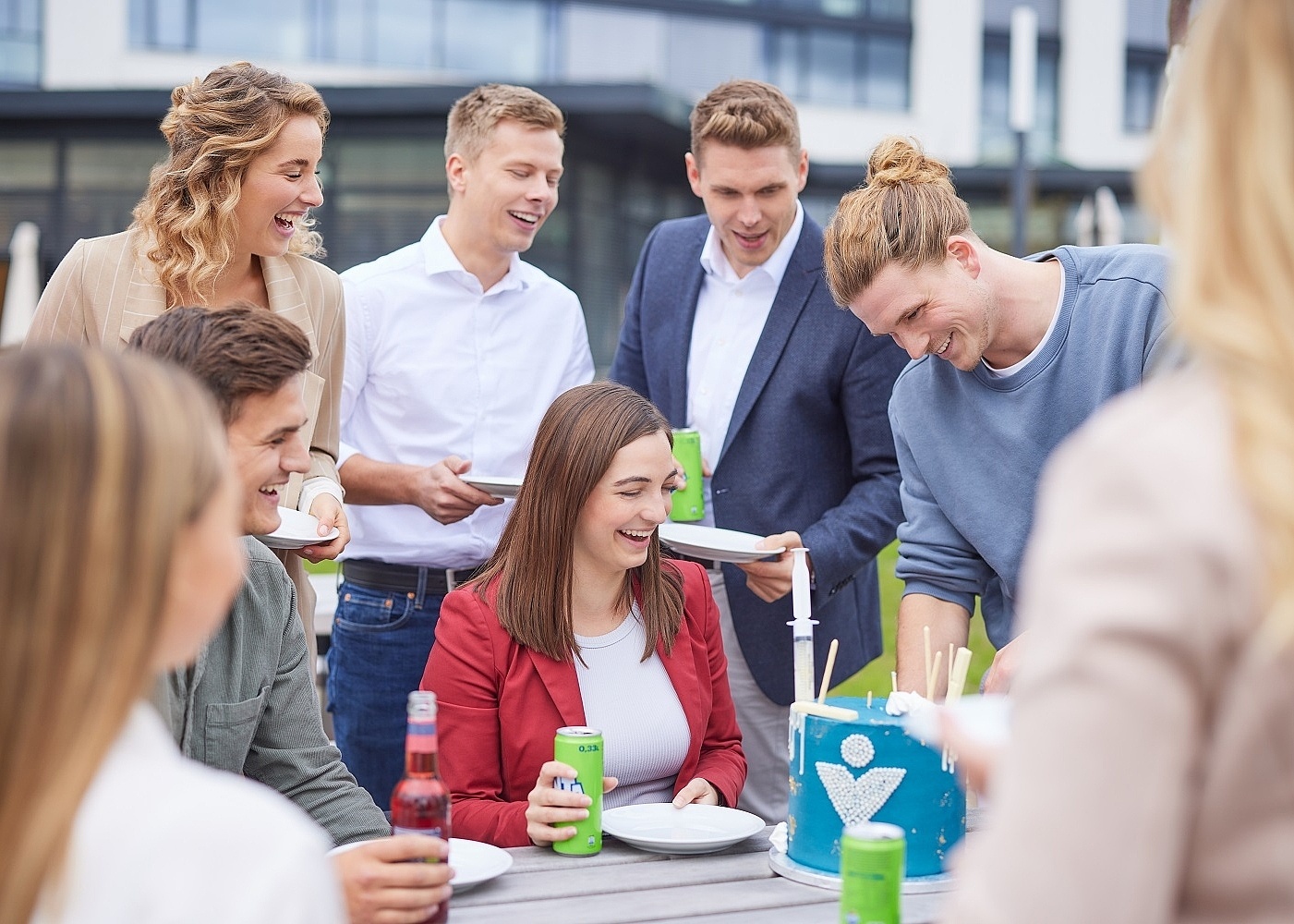 Lächelnde Mitarbeiter stehen und sitzen um einen Tisch und essen Kuchen 