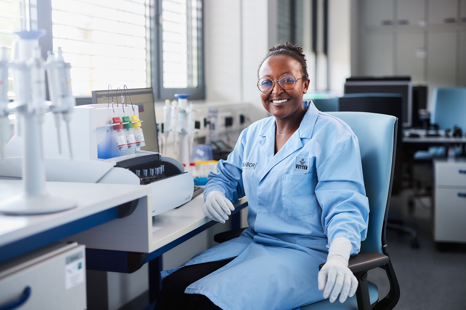 Eine Mitarbeiterin mit Laborkleidung sitzend auf einem Stuhl vor einer Maschine in der optischen Kontrolle