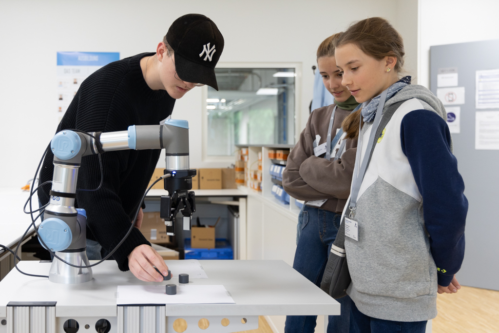 Ein Vetter-Azubi zeigt zwei Mädchen einen kollaborativen Roboter am Vetter Girls Day 2024