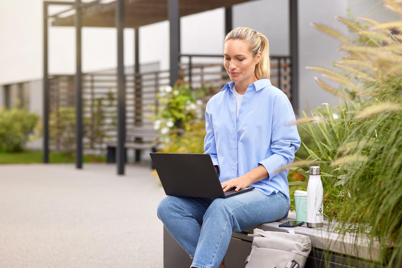 Eine Mitarbeiterin sitzt im Außenbereich auf einer Bank mit dem Laptop auf den Beinen und arbeitet im Freien